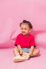 portrait of an 18 month old curly haired girl sitting laughing out loud while looking away isolated on pink