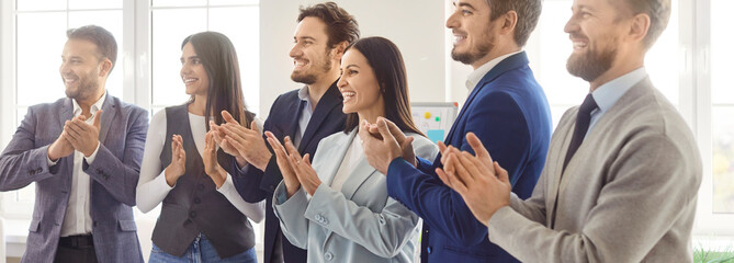 A group of six smiling professionals is clapping in a modern office setting. The diverse team showcases a sense of achievement during a corporate event.