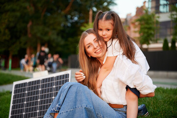 Naklejka premium Mother and daughter sit together on grass, embracing in warm moment with solar panel beside in the evening. Concept of renewable energy and family, highlighting commitment to sustainable future.