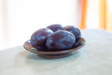 Four dark purple plums with drops of water on a brown plate on a light table