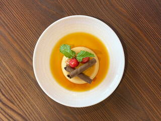 Vanilla butter pudding with maple syrup, chocolate sticks and cherry with mint leaves. Served on a white plate, isolated on wood table background. Dessert photography 