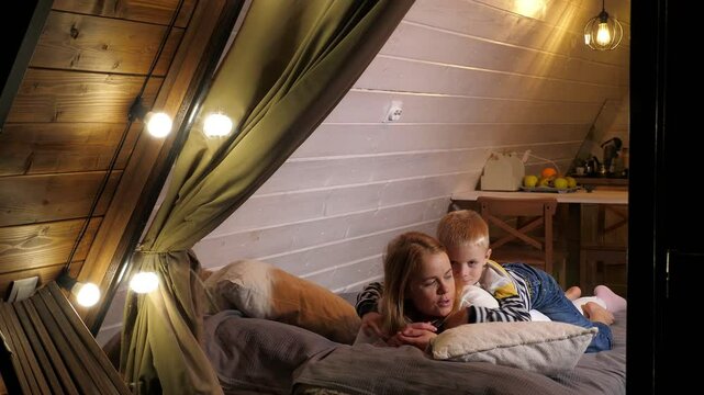 A mother and son in warm clothes are lying on a bed in a cozy wooden house in the forest in autumn. Family vacation in a small triangular house, view through the window in the evening.