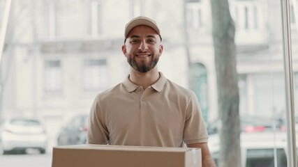Friendly courier man with a joyful expression delivering a large cardboard box, wearing a beige uniform and cap, showing satisfaction and confidence in his service, looking directly at the camera.
