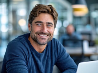 Obraz premium A smiling man sits in an office environment, exuding a sense of confidence and approachability. The background is slightly blurred, emphasizing a modern and professional setting.