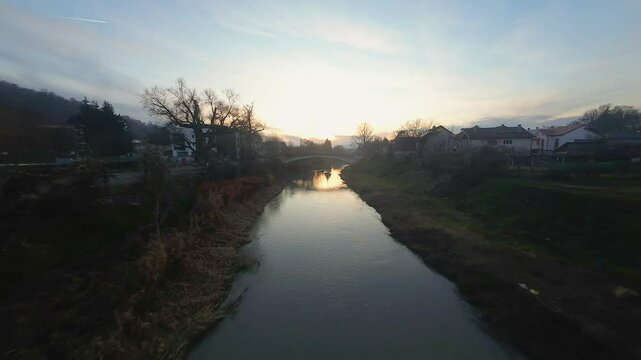Drone aerial view following a river and going under a bridge in the Town of Bistrita, Transylvania, Romania, East Europe during sunset, dusk capturing the city's outskirts.