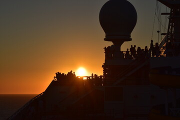 Obraz premium A stunning sunset over the ocean with silhouettes of passengers on a ship's deck, the warm glow of the sun creating a serene and tranquil scene as it sets behind the horizon 