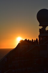 Obraz premium A stunning sunset over the ocean with silhouettes of passengers on a ship's deck, the warm glow of the sun highlighting the peaceful scene as it sets on the horizon, creating a serene atmosphere at se