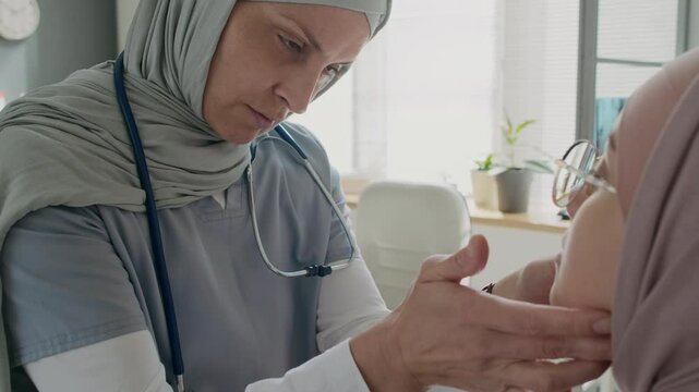 Close-up shot of female doctor in hijab warming her palms for performing lymph node palpation on Muslim patient sitting in medical office on examination couch