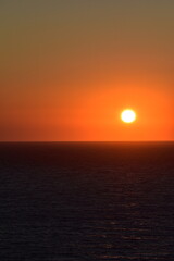 Sunset over the ocean, with a vibrant orange sun casting a warm glow across the calm water, and distant land visible on the horizon, creating a serene and peaceful seascape
