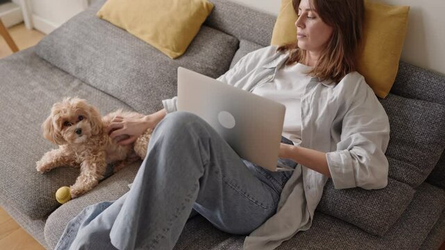 A woman multitasks on a comfortable sofa, working on a laptop while affectionately petting her cute Maltipoo dog beside her.