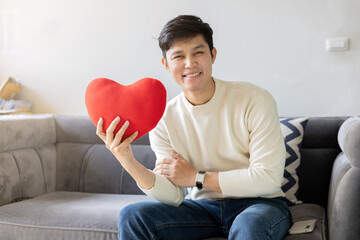 close up young asian man smile with happiness feeling and hold red color of heart shape pillow while sit on couch in living room for good health and live better and valentine day and people lifestyle