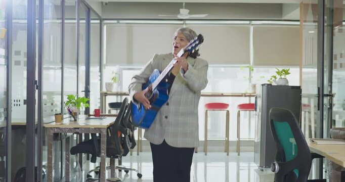 Indian Asian happy senior gen x business woman play guitar singing song indoor job place excited cute old silly crazy lady staff worker dancing enjoy day break time have fun joyful joy work office