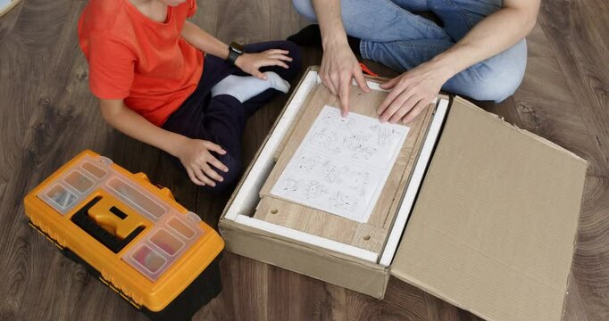 Father and son sit on the floor, unpacking furniture while studying the instruction manual. They share a moment of teamwork and learning, surrounded by boxes and tools