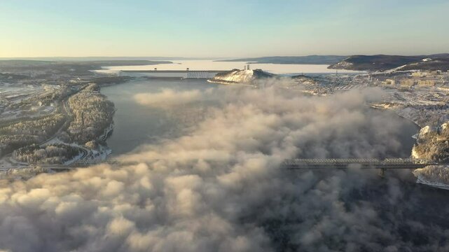 Drone flight at sunset over a lake with a dam in winter. The fog over the lake is illuminated by the setting sun. The dam is near the city.