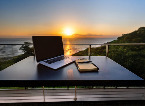 Laptop and notebook sit on a table overlooking a stunning sunset.  A tranquil home office or remote work setup with ocean views.