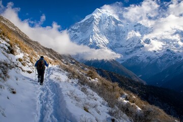 A hiker climbs a snow-lined path toward a towering mountain peak, embraced by clouds, capturing the magnificent allure and adventurous spirit of high-altitude exploration.