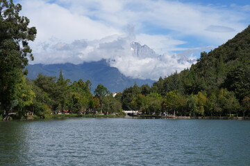 Majestic snow-capped mountains rise above a serene lake, surrounded by lush trees. The peaks are partially veiled by soft clouds, while the crystal-clear lake mirrors the breathtaking scenery.