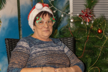 A smiling woman wearing a Santa hat and decorative glasses with holly accents sits near a Christmas...