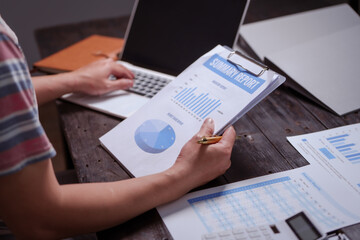 A man sits at his desk at home at night, working on his laptop. He views charts, checks documents,...