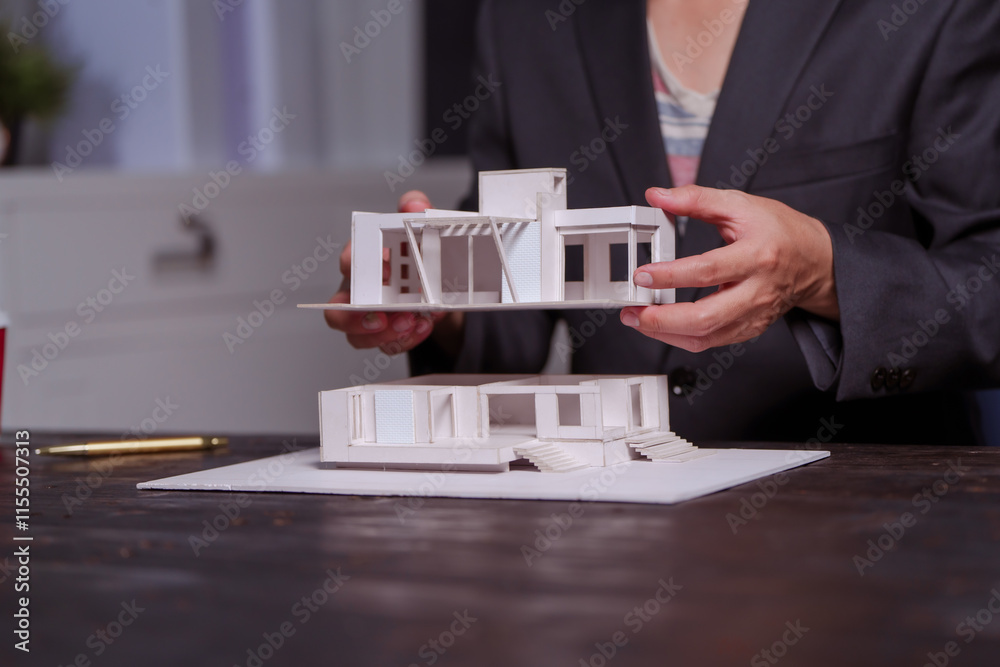 Wall mural A businessman sits at a table at night, holding a model of a two-story white house. He reviews designs, drafts plans, and inspects the roof details, showcasing modern architecture