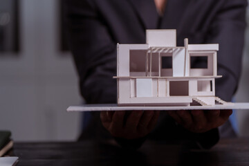 A businessman sits at a table at night, holding a model of a two-story white house. He reviews...