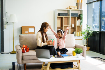 An Asian mother and her adorable daughter sit on the sofa in their living room, enjoying online learning on a tablet and reading books. A joyful family moment focused on education