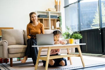 An Asian mother and her adorable daughter sit on the sofa in their living room, enjoying online learning on a tablet and reading books. A joyful family moment focused on education