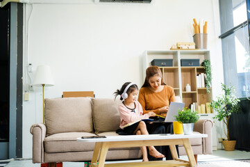 An Asian mother and her adorable daughter sit on the sofa in their living room, enjoying online...