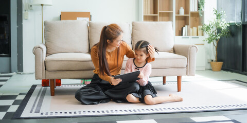 An Asian mother and her adorable daughter sit on the sofa in their living room, enjoying online...