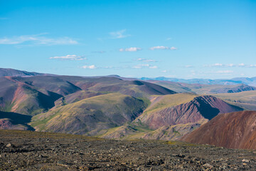 Scenic top view from sunlit stony plain to big colorful mountain range in bright sun under clouds in blue sky. Layered alpine scenery with large multicolor mountains of many vivid colors in sunny day.