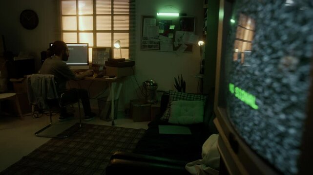 Man with beard sitting at desk, working on computer and drinking from mug in dimly lit room with green neon lights