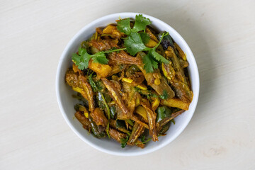 Mola shutki bhuna, a traditional Bengali dry fish curry, is served in a white bowl on a light wooden surface. It is deliciously cooked with potato and brinjal. Top view.
