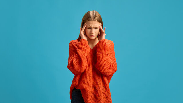 Young woman in orange sweater holding her temples, expressing discomfort from headache isolated on blue background. Tension headache. Concept of illness, medicine, treatment, health care