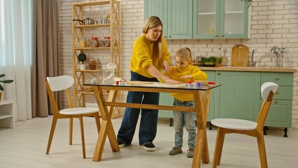 Mother and daughter together in the kitchen, mom teaching girl how to make gingerbread cookies, mom helps the little girl to roll the dough with rolling pin. Full shot.