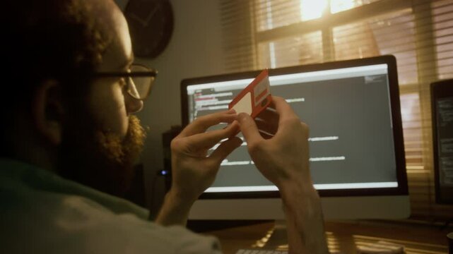 Bearded man examining and playing around with red floppy disk at desk with computer screen displaying code in sunlit room