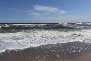 Waves with white foam washing onto sandy shore under a clear blue sky. Coastal seascape photography. Nature and ocean concept for design and print.