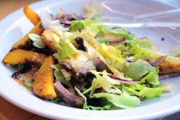 a portion of food from green fresh salad with fried potatoes and meat on a white plate on a table in a room in a restaurant