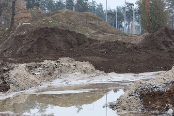 big piles of black grey earth and white sand on construction site with puddle on road on autumn street