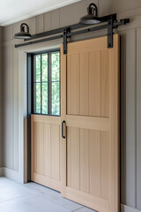 Fototapeta premium Barn door with black metal sliding vertical track, in a light wood color. In the background, there is an open window and wall sconces on either side of it. The room has white walls with gray trim.