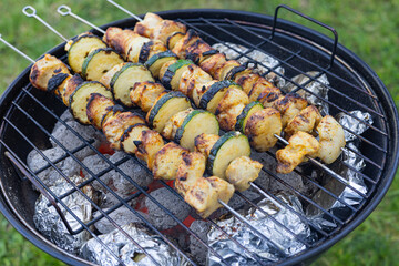 Grilled chicken and zucchini skewers cooking on a charcoal barbecue with baked potatoes wrapped in foil. Outdoor cooking, summer picnic and delicious barbecue food in a backyard setting