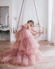 young brunette girl in pink princess dress with train is sitting on the crystal chandelier near...