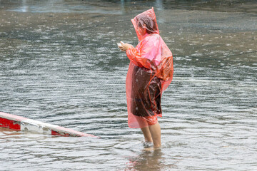 A woman in a raincoat stands in the middle of a large puddle and looks at her mobile phone