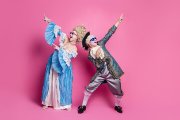Playful couple dressed in extravagant period costumes strikes playful pose against vibrant pink backdrop, embodying historical elegance