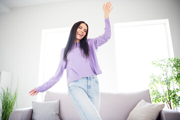 Young brunette woman joyfully dancing in living room with daylight streaming through windows, wearing a stylish purple pullover