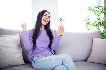 Obraz premium Smiling young woman enjoying a joyful moment at home using a smartphone, sitting comfortably on a stylish couch