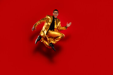 Energetic young man in a glossy gold suit jumps joyfully against a vivid red backdrop, embodying fun and style.