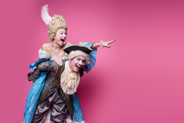 A joyful couple dressed in elegant historical costumes enjoys a playful moment against a vibrant pink background