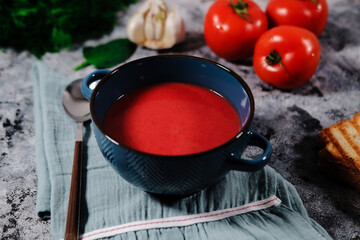 beetroot or tomato soup served in dark blue bowl, kitchen cloth on gray background, fresh tomatoes, garlic, roasted slice of bread