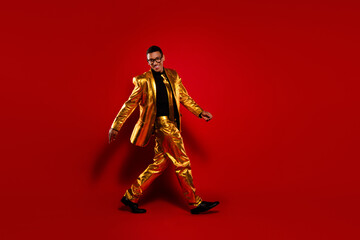 Charismatic young man in a glossy golden suit walking against a vibrant red background, embodying a sense of celebration and style