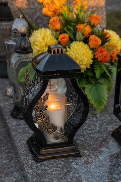 A beautiful candle shines on the grave, in the background yellow chrysanthemums and orange roses.